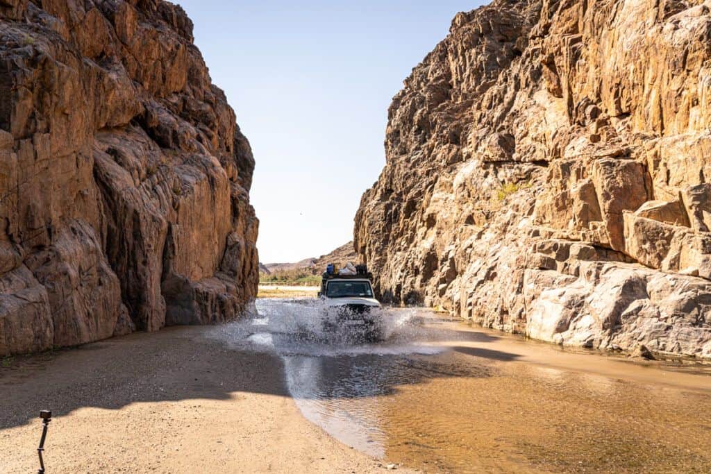A 4x4 driving through a canyon, through a pool of water with spray ahead of the vehicle