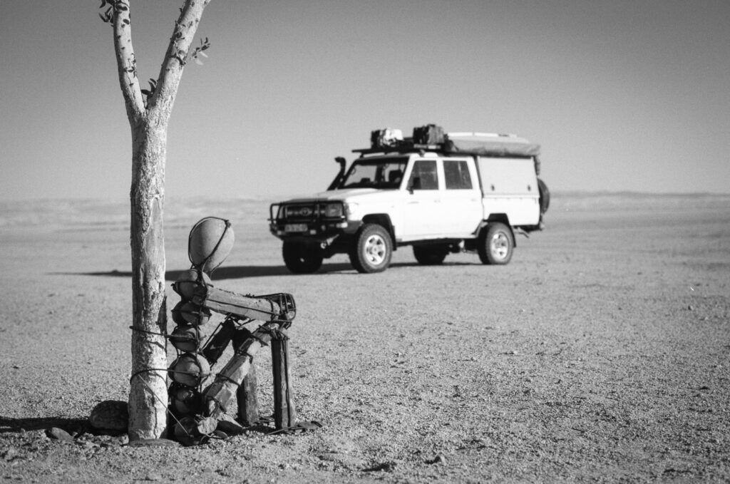 A black and white image of a stone man of Kaokoland sitting by a tree with a 4x4 in the background